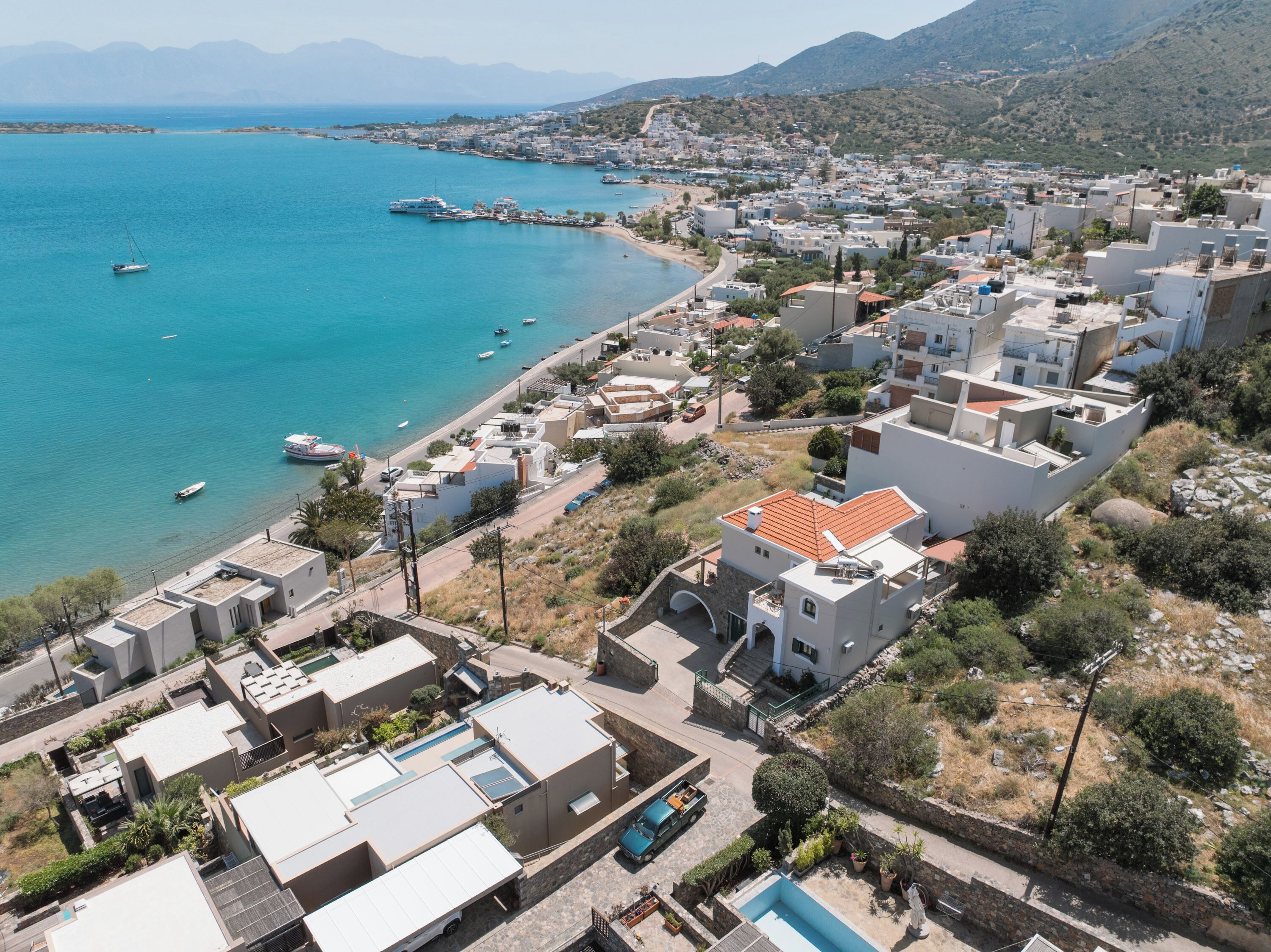 A coastal town with modern buildings, a clear blue sea, and a mountainous backdrop.