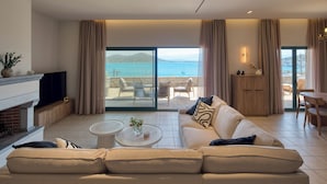 Een moderne woonkamer met een bank, salontafel en een open haard. Er is een balkon met uitzicht op zee en bergen.