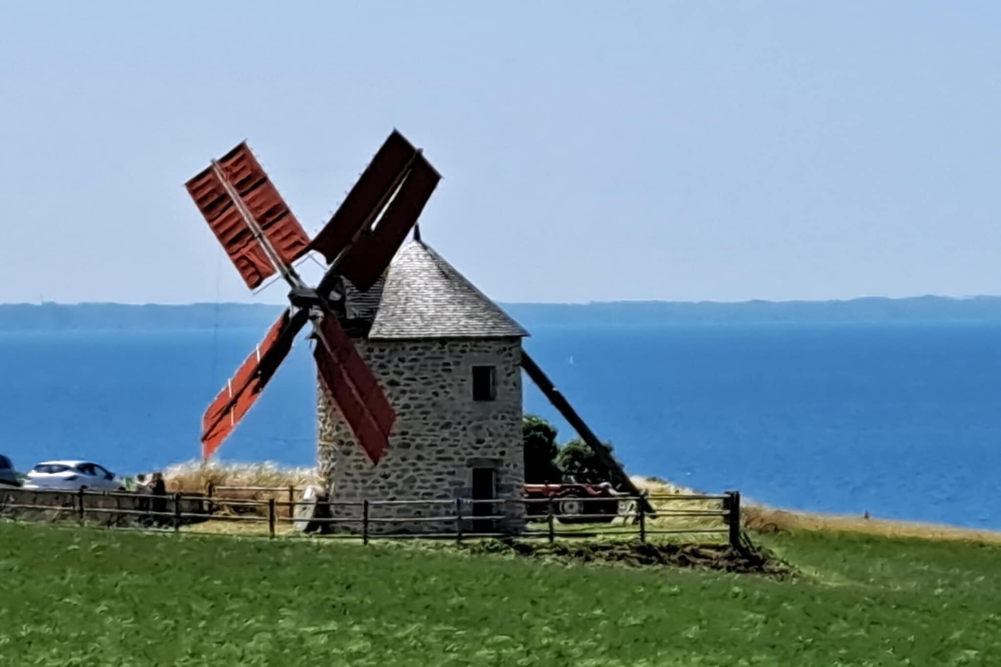 A stone windmill with a red windlass, set against a backdrop of a clear blue sea and distant hills.