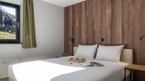 A modern bedroom with a large bed, a window with a view of a house, a wooden accent wall, and a bedside table with a lamp. - Villa in Belgium with Private Terrace (La Roche-en-Ardenne)