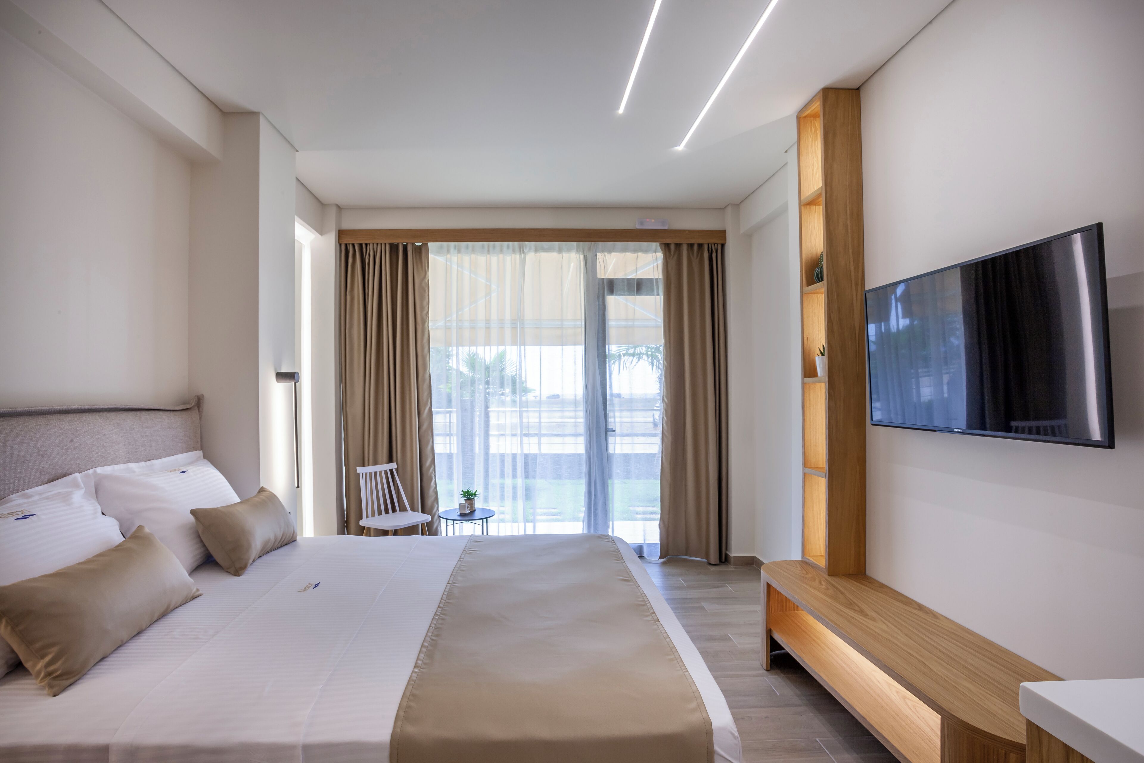A modern hotel room with a large bed, a TV mounted on the wall, and a view of the outdoors through the window.