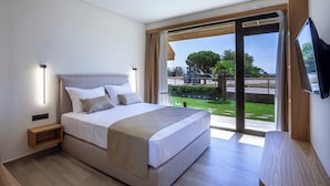 Ein modernes Schlafzimmer mit einem großen Bett, einem Nachttisch, einem Fernseher und einer Schiebetür zu einem Balkon mit Blick auf Grünflächen und eine Straße.