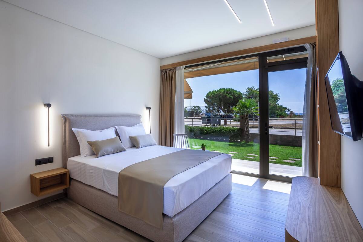 A modern bedroom with a large bed, a nightstand, a TV, and a sliding glass door leading to a balcony with a view of greenery and a road.