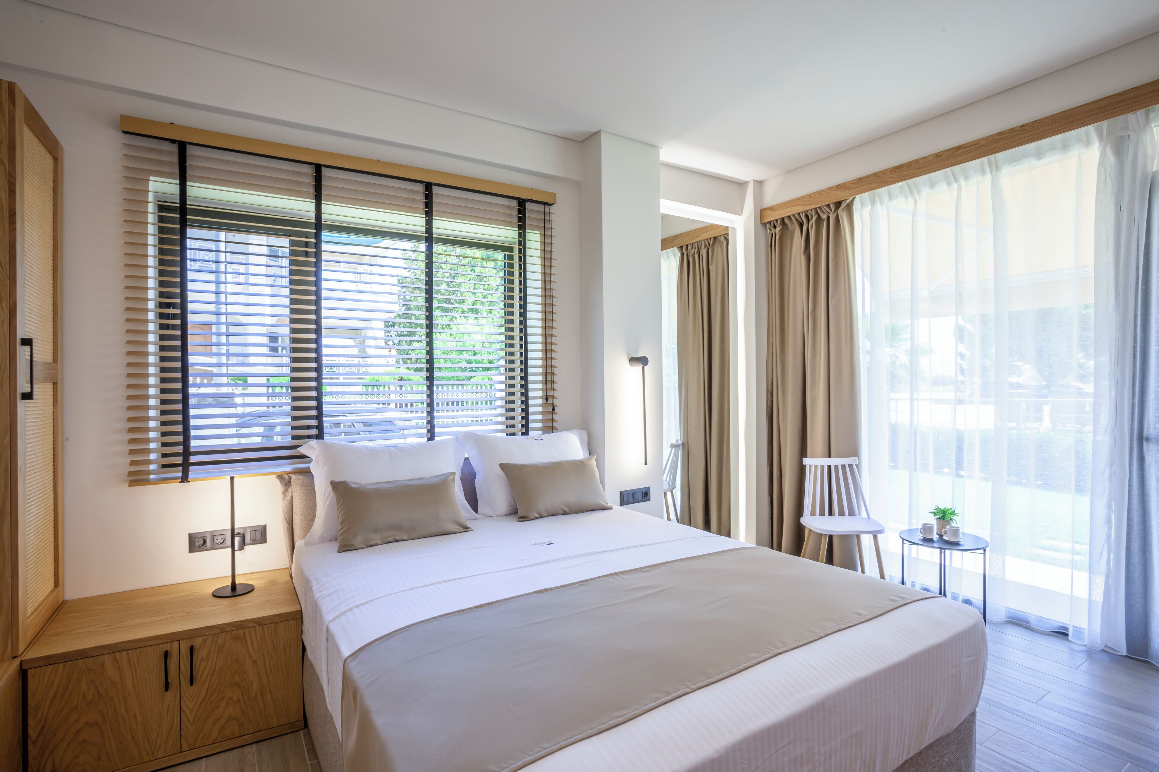 Una habitación de hotel moderna con una cama grande, una mesita de noche, una lámpara, una pequeña mesa con una planta y vistas al exterior a través de amplios ventanales.