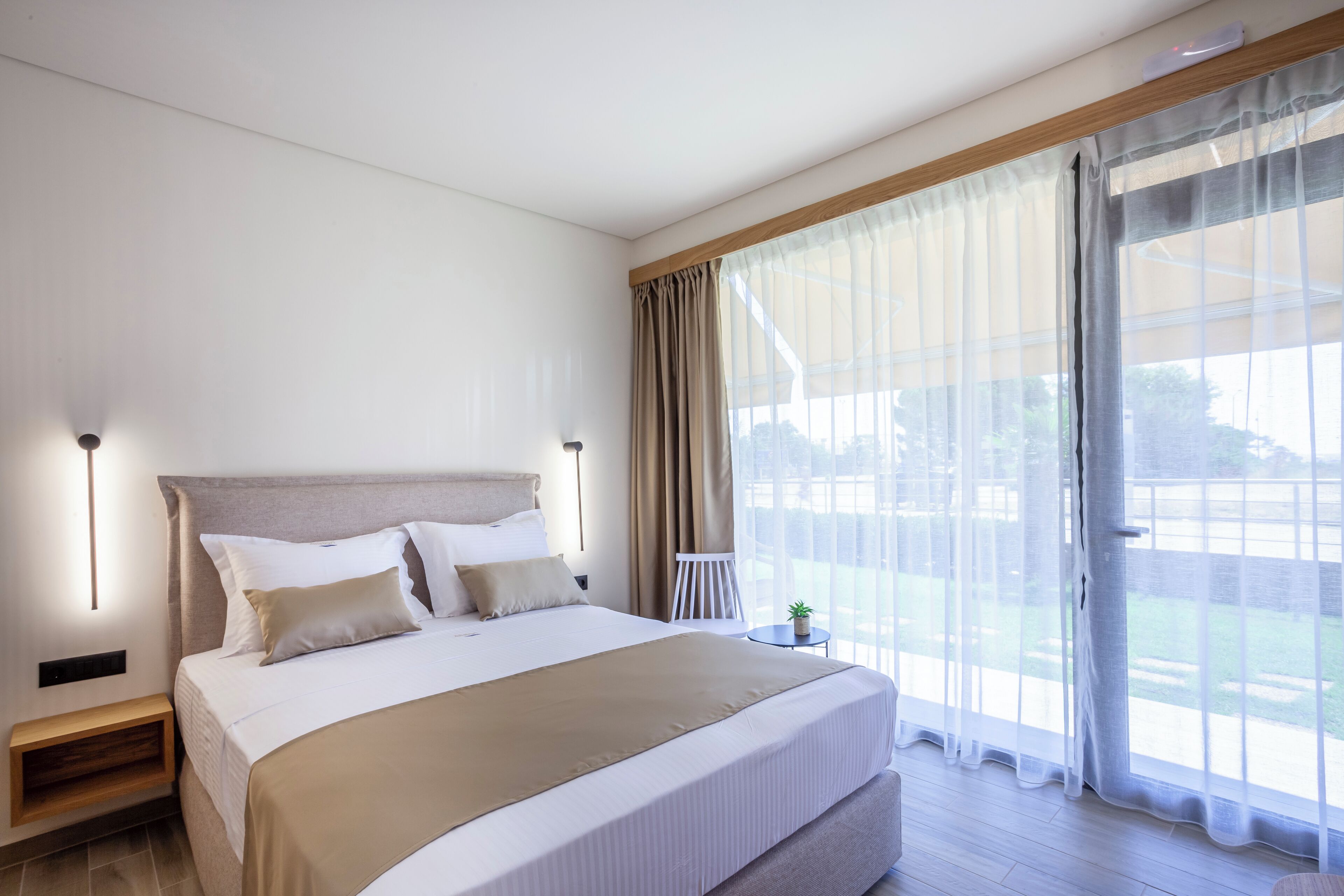 A modern hotel room with a large bed, a bedside table, a small table with a plant, and a view of the outdoors through sheer curtains.