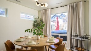 A dining area with a round table set for four, surrounded by brown chairs. There is a potted plant on the table, and a metal cart with glasses and a bowl on the right.