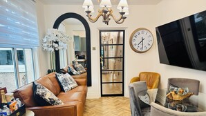 A modern living room with a brown leather sofa, a grey armchair, a glass coffee table, a large wall clock, and a flat-screen TV. - Luxury 2 Bed Designer Flat with Private Garden in London (Bexleyheath)