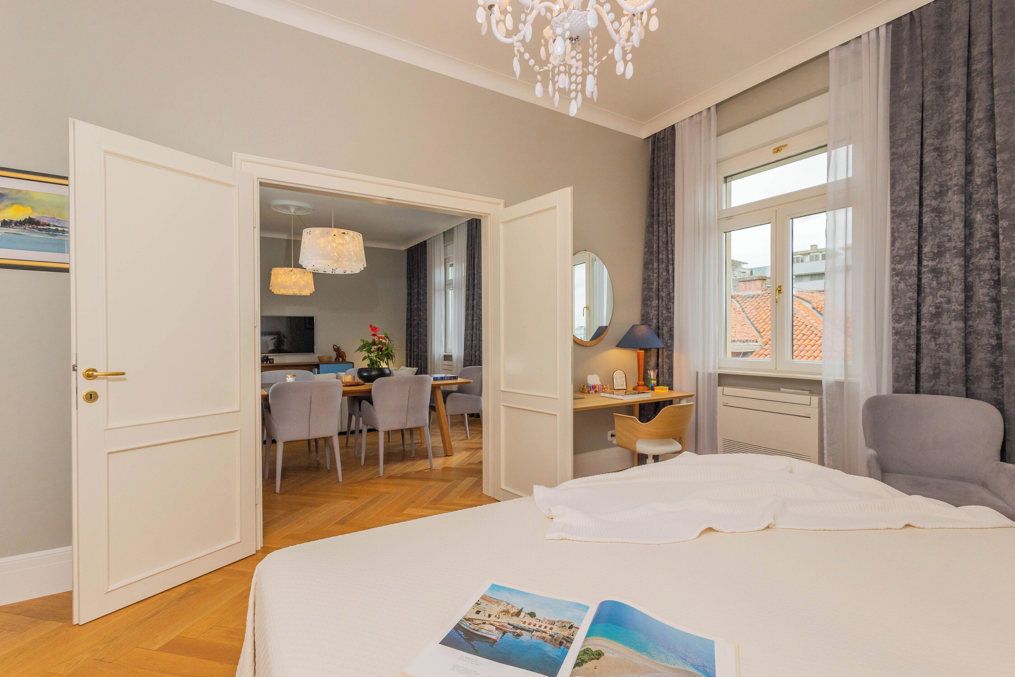 Et hotelværelse med en seng, et skrivebord, en stol, en lysekrone og udsigt over byen gennem vinduet.