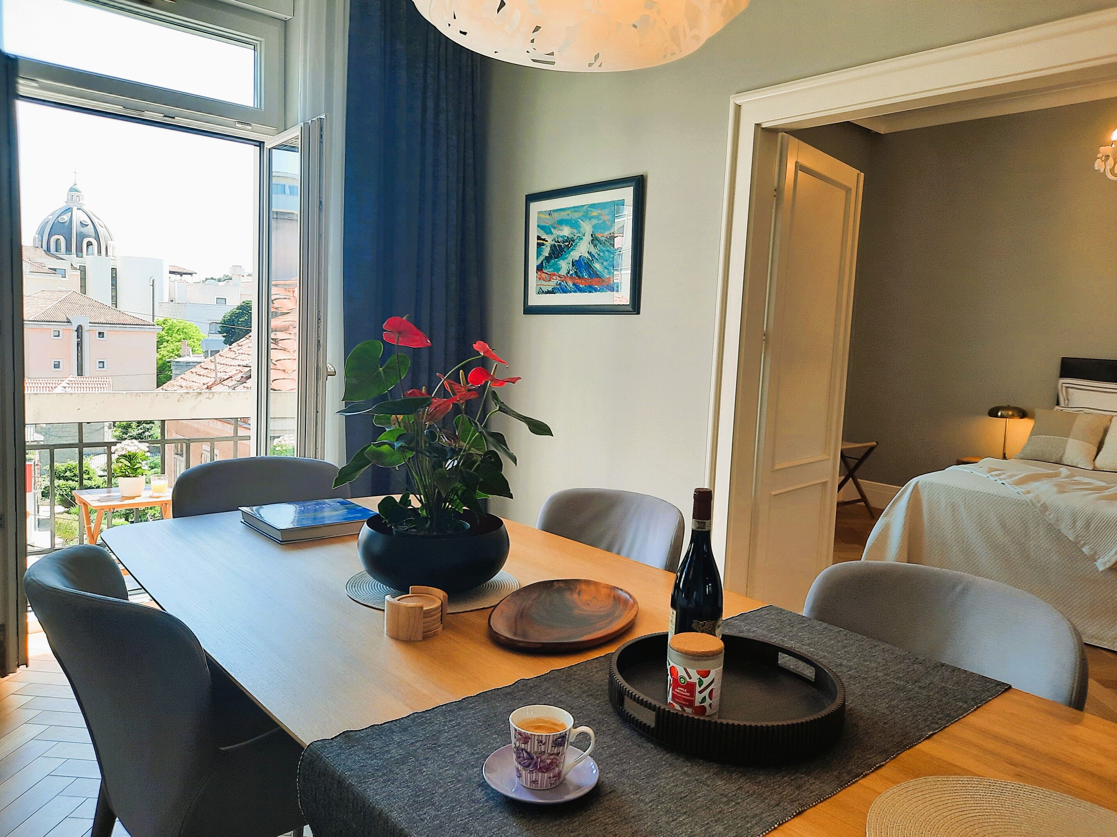 A hotel room with a dining table, chairs, a potted plant, a bottle of wine, and a bed.