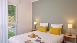 A neatly made bed with white and yellow pillows, a straw hat, and sunglasses on the bed. There are two bedside lamps on wooden nightstands.