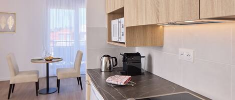 A modern kitchen with a built-in microwave, coffee maker, and induction cooktop. There is a dining area with a table and chairs, and a view of the outside through a window.
