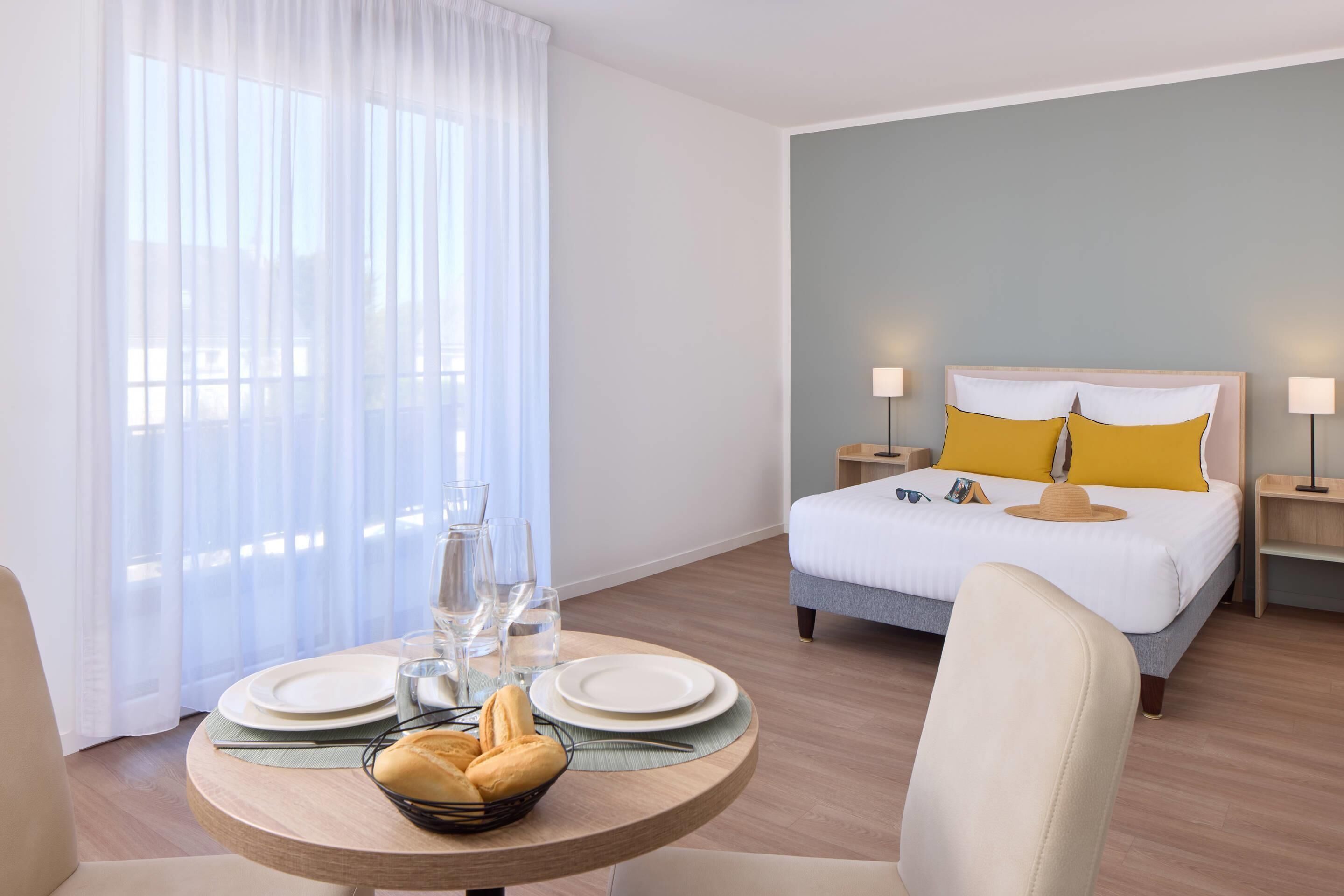 Een hotelkamer met een bed, bedlampen, een eettafel met borden en glazen, en een raam met gordijnen.