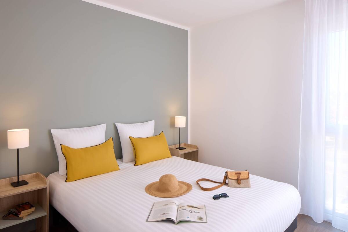 Un lit bien fait, avec du linge de lit blanc et des oreillers jaunes, un chapeau, des lunettes de soleil et un livre posés dessus. Il y a deux tables de chevet avec des lampes et une petite étagère remplie de livres.