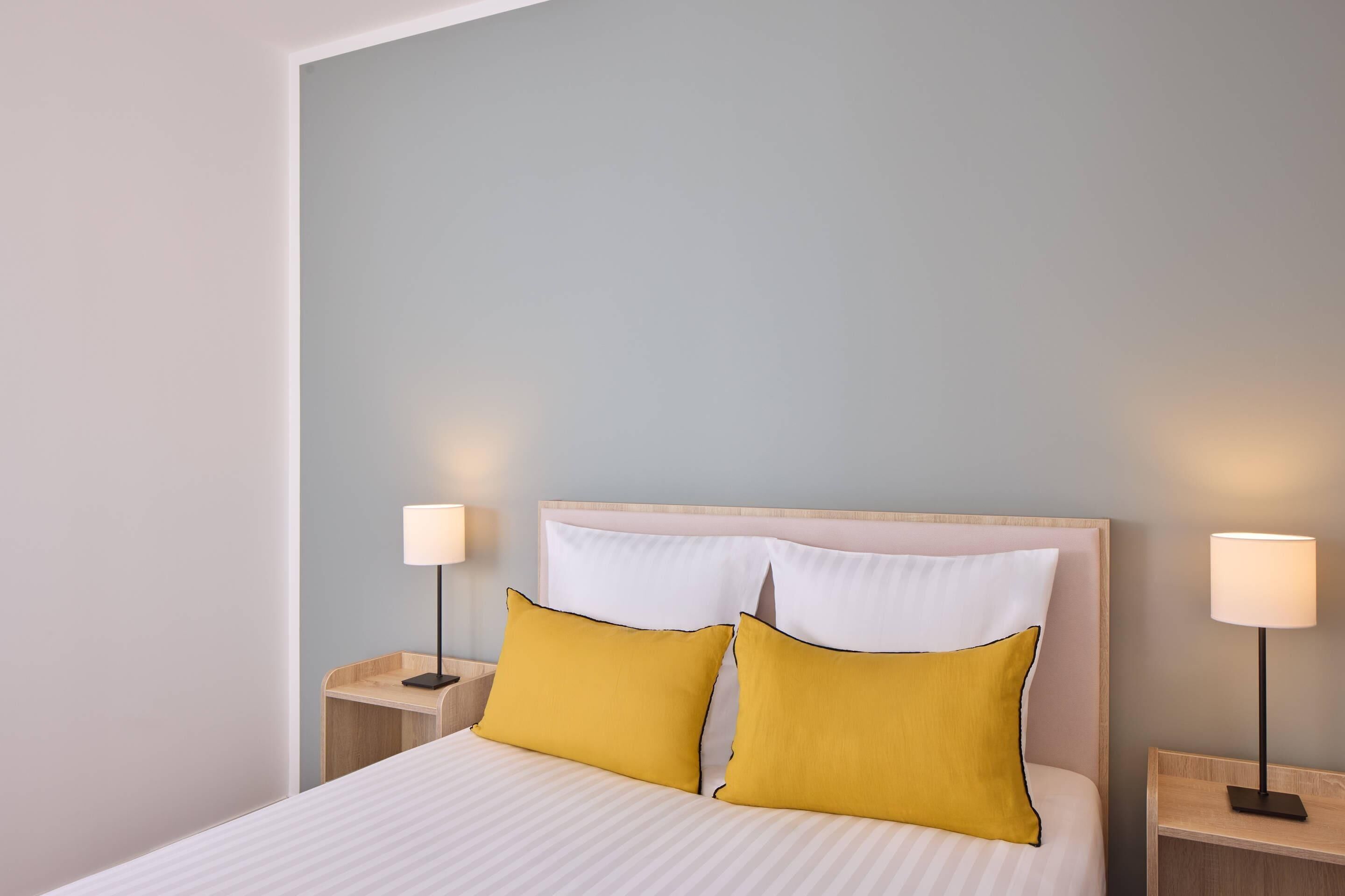 A neatly made bed with white bedding and two yellow pillows, flanked by two bedside tables with lamps, in a room with light blue walls.