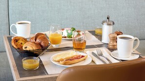 A breakfast tray with croissants, orange juice, and a coffee cup on a cushioned seat with a scenic mural in the background.
