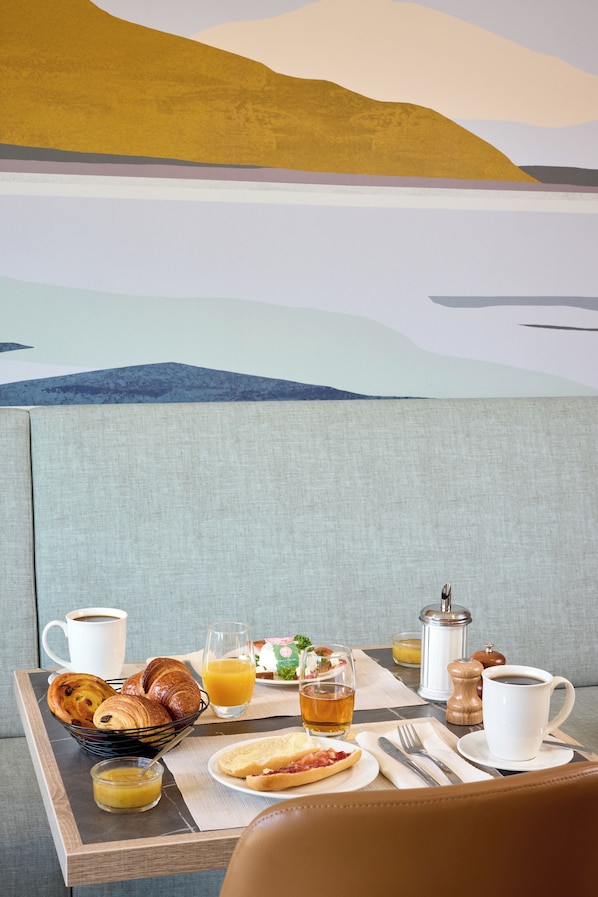 Bandeja de desayuno con cruasanes, jugo de naranja y una taza de café sobre un asiento acolchado, con un mural panorámico de fondo.