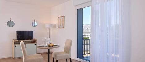 A hotel room with a bed, a desk with a TV, two chairs, a table with a lamp, and a view of the outside.