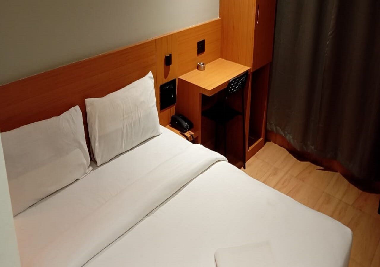 A hotel room with a wooden headboard, a single bed with white linens, a small wooden table with a lamp, and a dark curtain on the window.