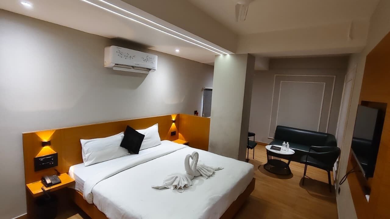 A hotel room with a neatly made bed, a bedside table with a lamp, a desk with a chair, and a TV.