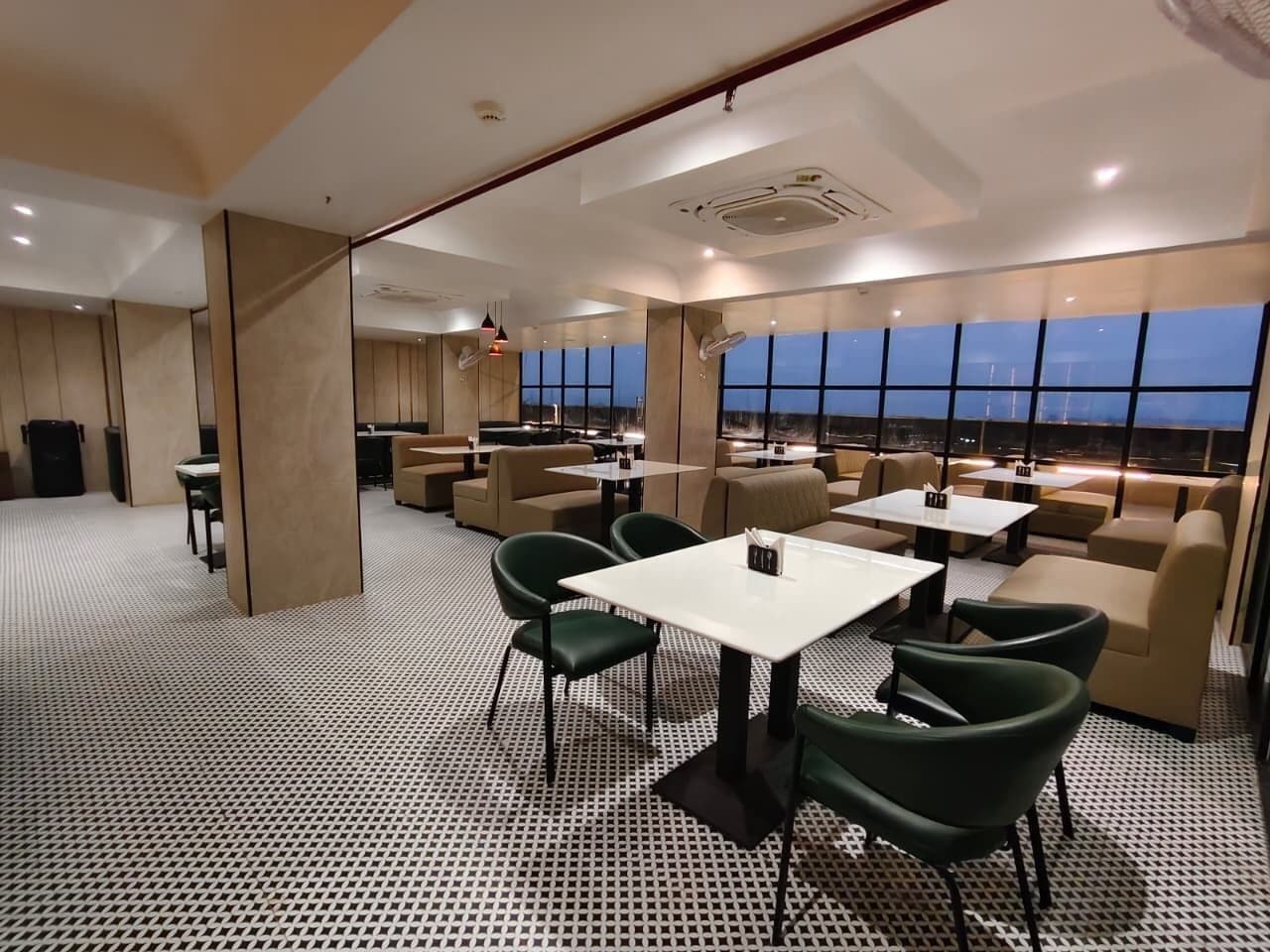 Un área de descanso moderna con piso estampado, asientos verdes y beige, y ventanas amplias que ofrecen vistas de la ciudad.