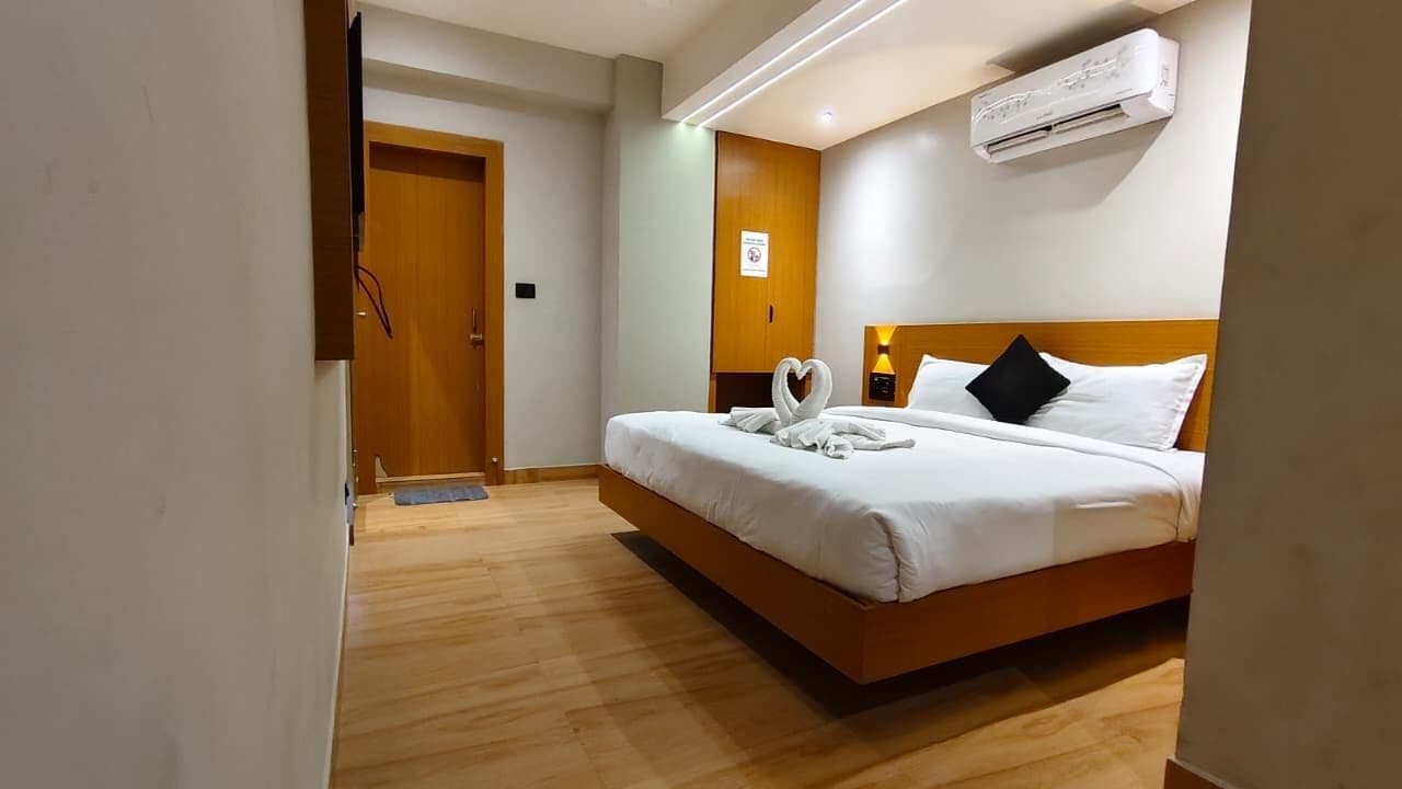 A hotel room with a wooden floor, a bed with white bedding, a nightstand with a lamp, and an air conditioning unit on the wall.
