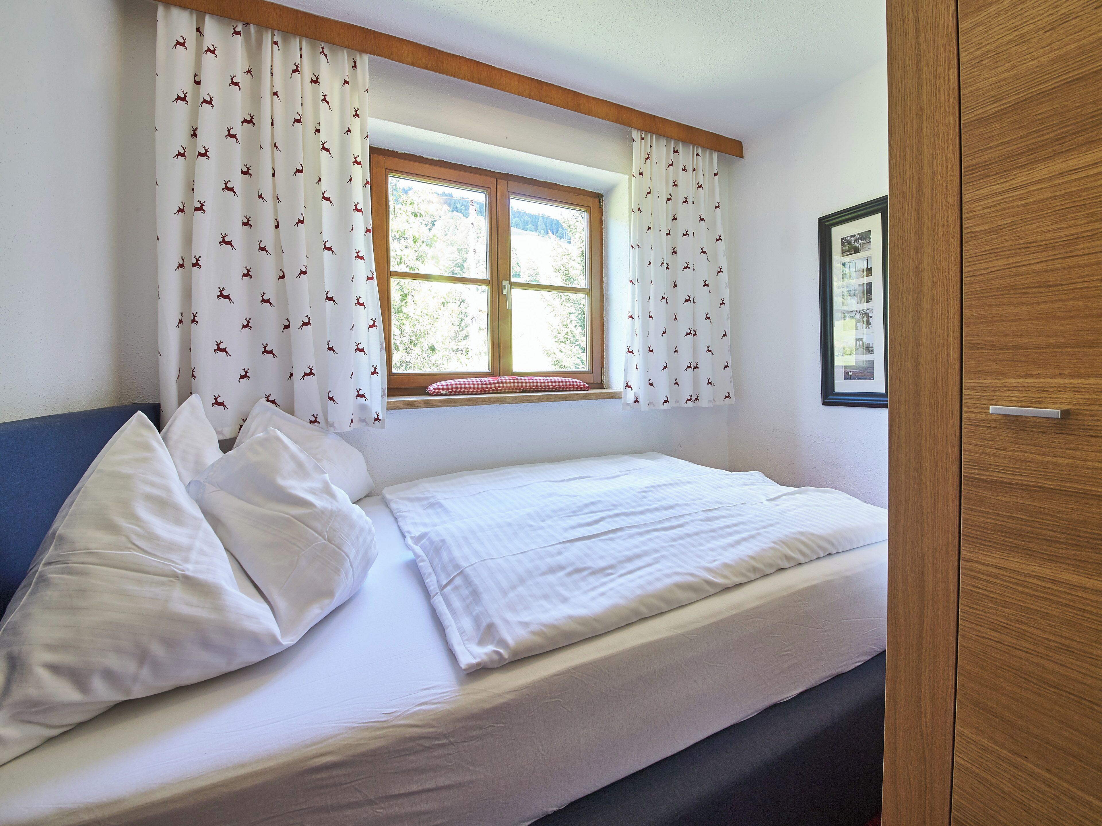 Une chambre à coucher avec un lit, des rideaux blancs ornés d’un motif de cerf, une fenêtre donnant sur la verdure et une armoire en bois.