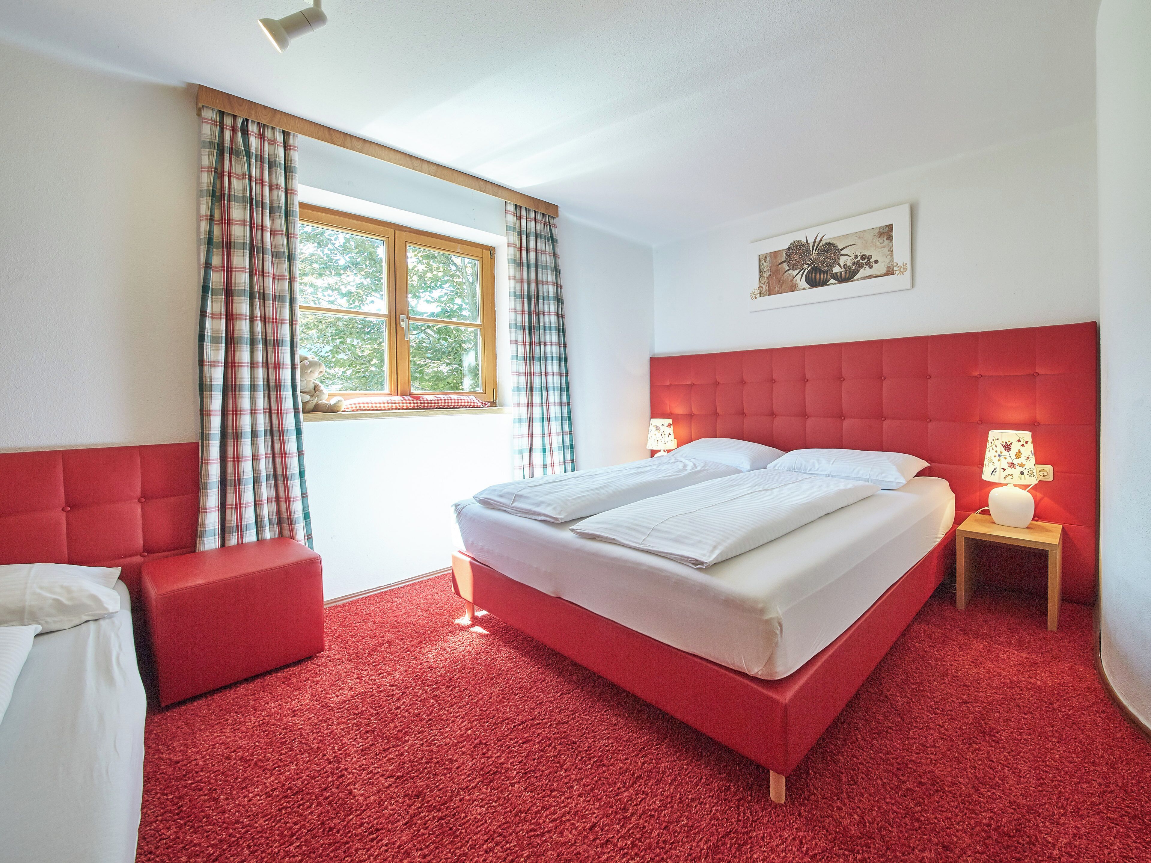 A bedroom with a red upholstered headboard, a large window with plaid curtains, a bedside table with a lamp, and a red carpeted floor.