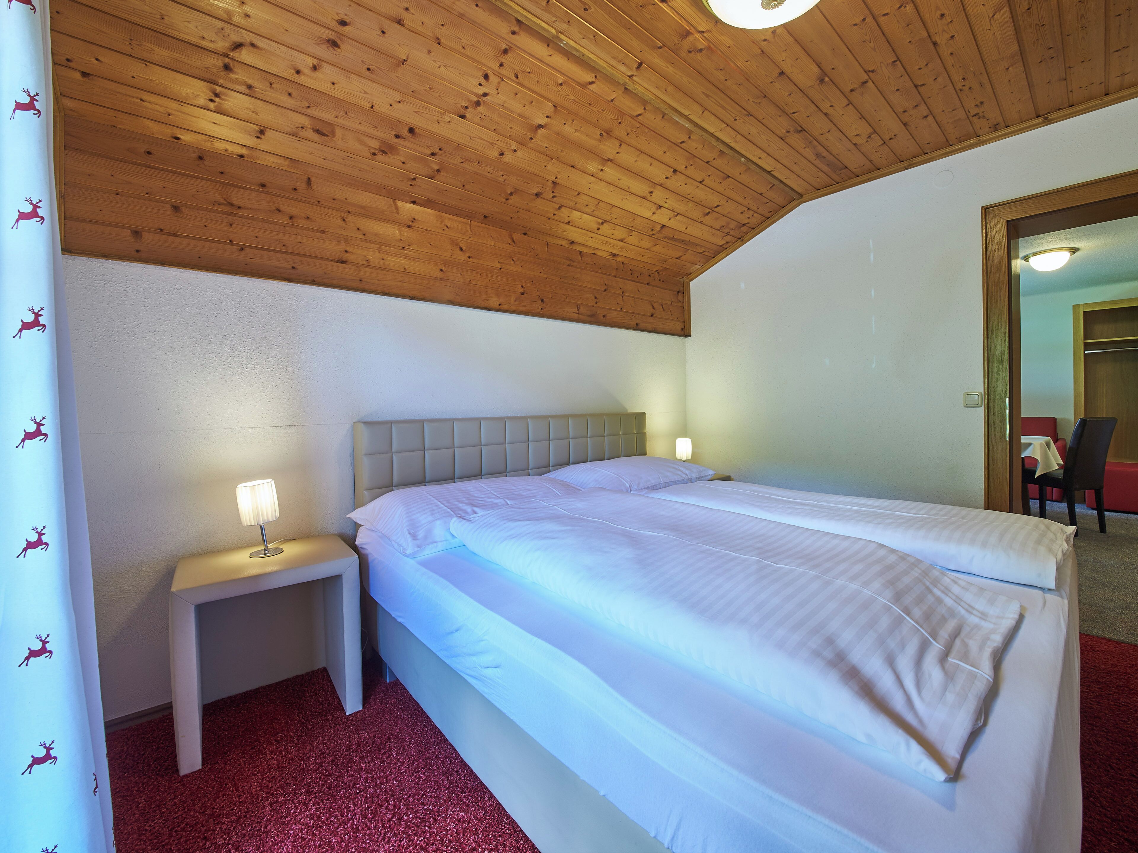 Quarto com teto de madeira, cama com roupa de cama branca, mesa de cabeceira com abajur e piso coberto por carpete vermelho.