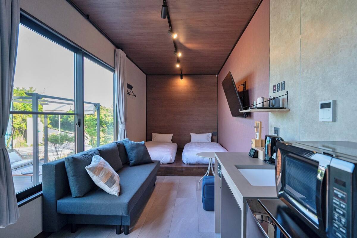 Una habitación de hotel moderna con sofá, dos camas, televisor y kitchenette.
