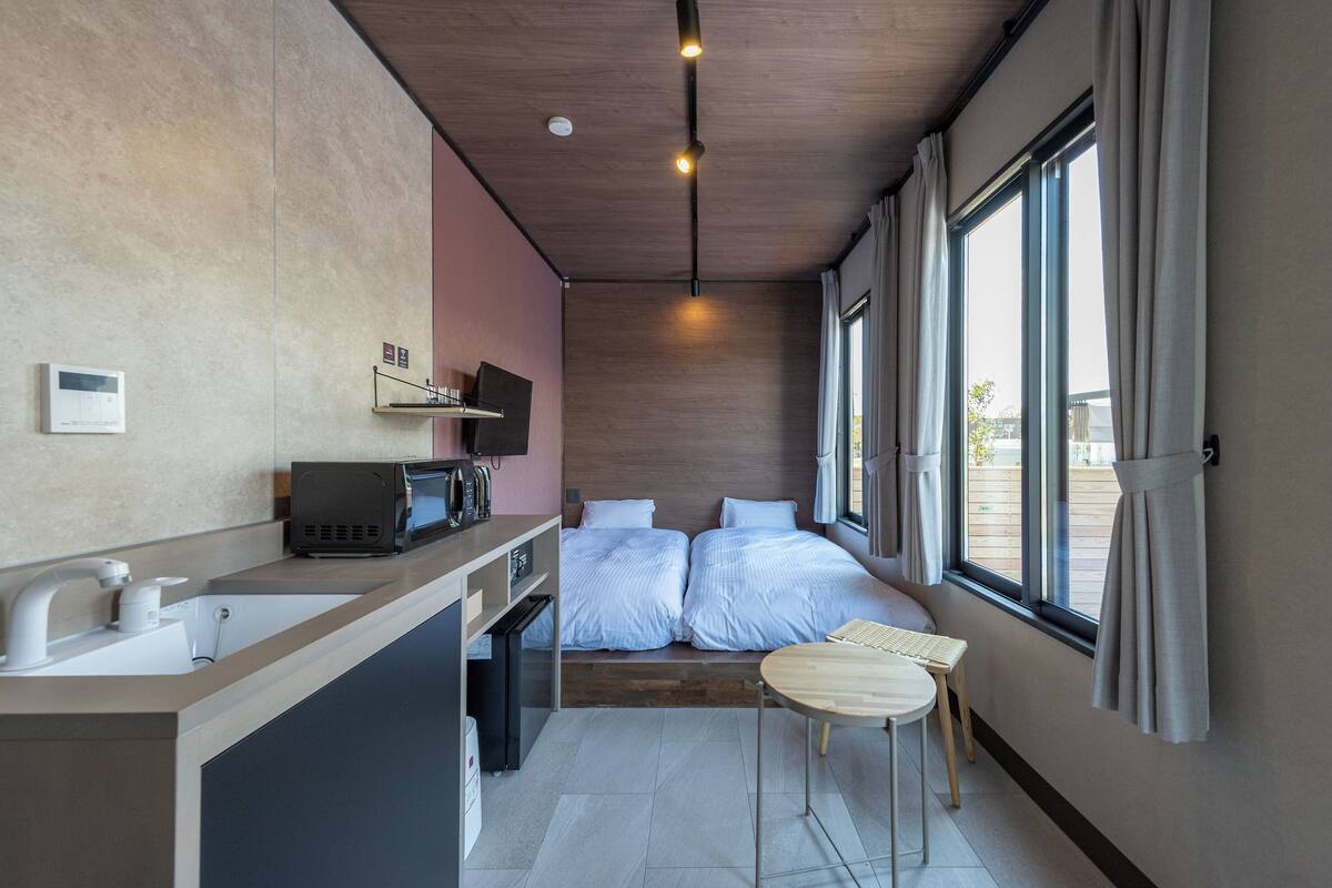 Una habitación de hotel moderna con techo de madera, cama con ropa de cama blanca, una pequeña mesa de madera, un lavamanos con espejo, un microondas y un televisor en un estante.