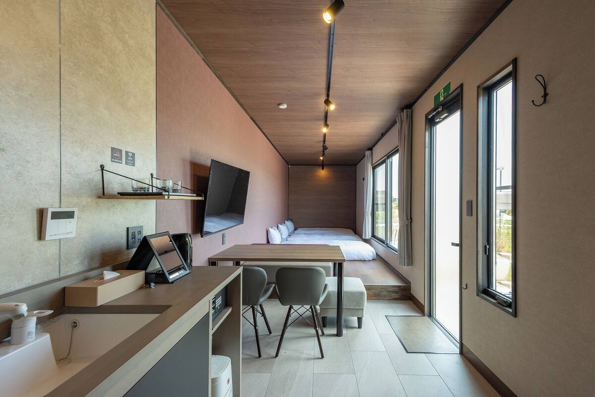 Una habitación de hotel moderna con un escritorio de madera, dos sillas, una cama con vistas, un televisor montado y una ventana con cortina.