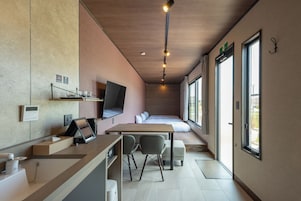 A modern hotel room with a wooden desk, two chairs, a bed with a view, a mounted TV, and a window with a curtain.