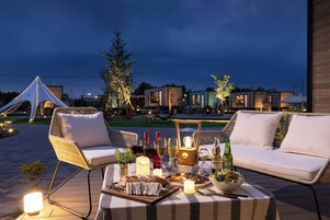 An outdoor dining area with wicker chairs, a table set with food, drinks, and candles, and a view of modern buildings in the background.