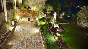 A well-lit outdoor area with a paved walkway, a seating area, and a small garden with a lawn and trees.