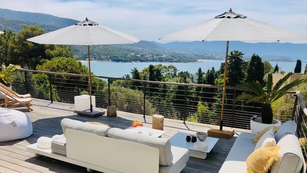 A deck with white sofas and two umbrellas, overlooking a lake and hills.