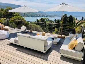 Una terrazza con divani bianchi e due ombrelloni, con vista su un lago e delle colline.