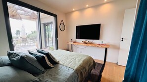 A bedroom with a bed, a TV, a wooden shelf, and a view of the outdoors.
