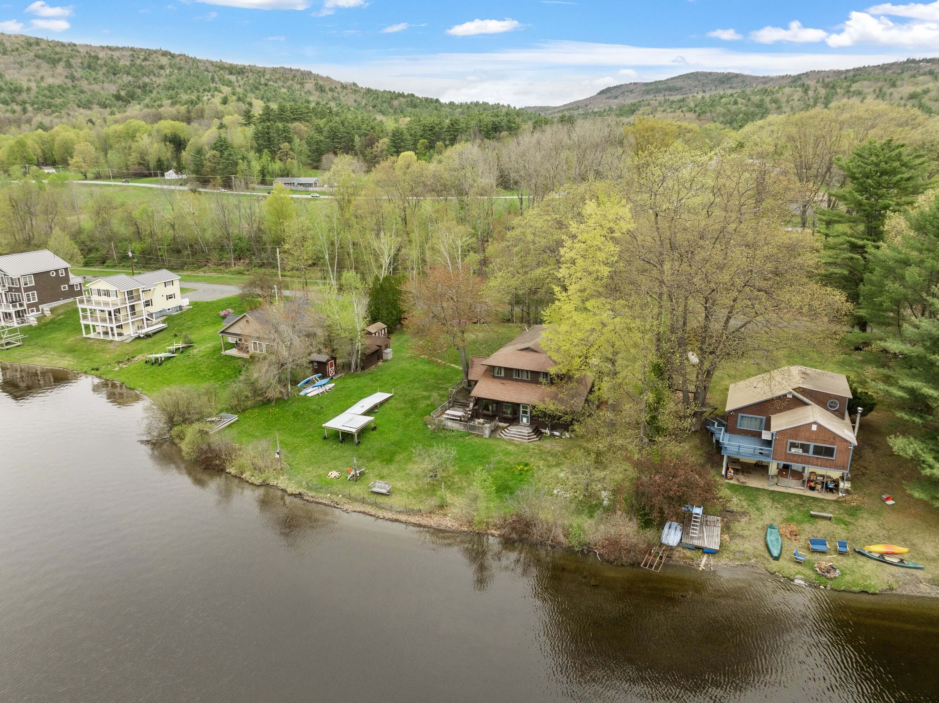 A lakeside property with multiple buildings, including a large multi-story house and smaller structures, surrounded by trees and a hilly landscape.
