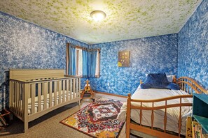 Ein Schlafzimmer mit einem Kinderbett, einem Bett mit blauem Kopfkissen, einem farbenfrohen Teppich und einer Decke mit einer Leuchte.