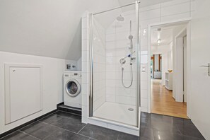 A modern bathroom with a glass shower enclosure, a washing machine, and a visible hallway leading to a living area.