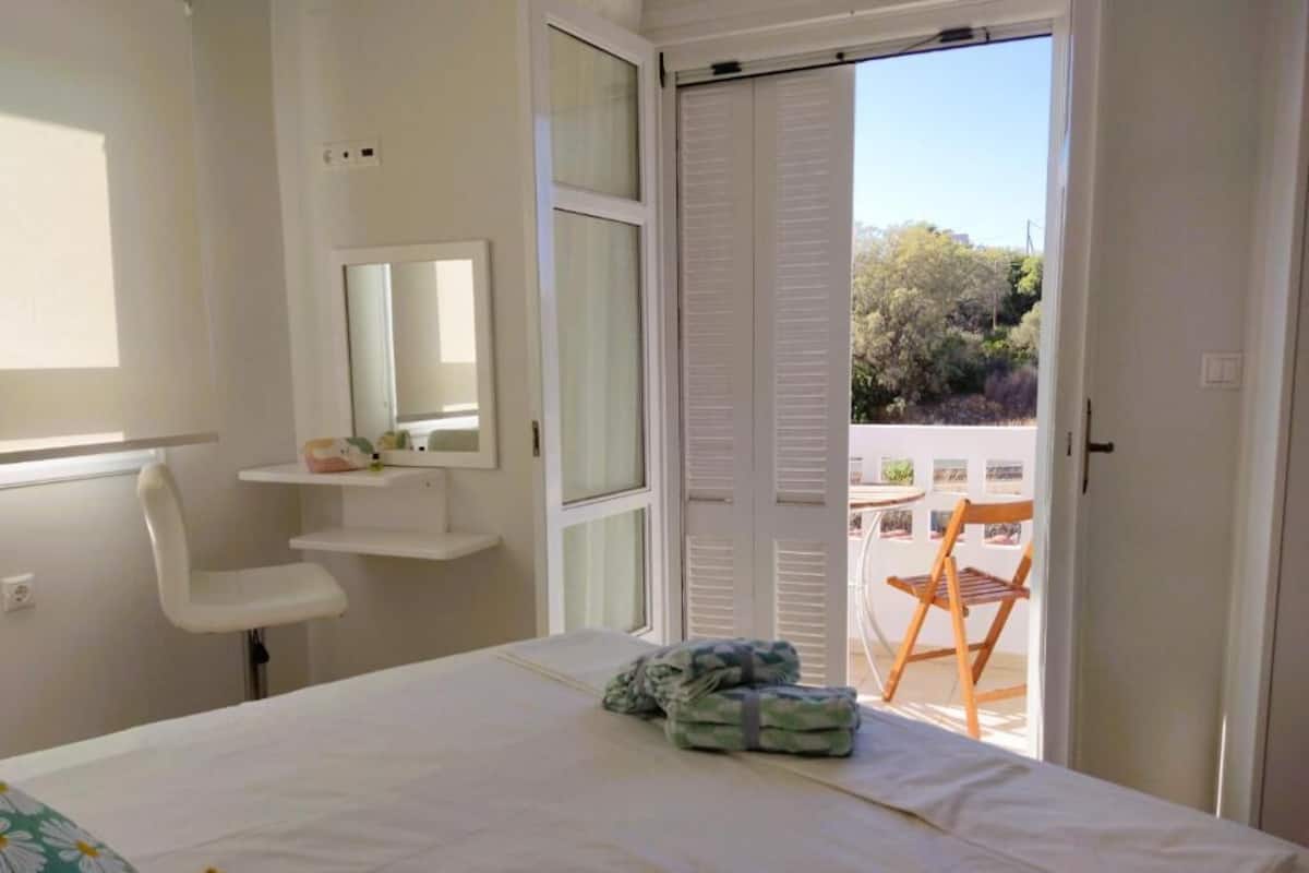Un dormitorio con cama, una silla, una mesita, un espejo y una puerta corrediza que da a un área exterior con una silla y una mesa.
