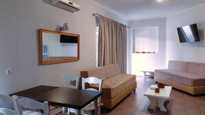 A living room with a sofa, a dining table with chairs, a TV mounted on the wall, and an air conditioning unit. - Tarmaros - Families & Groups (Sissi)