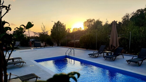 A rectangular swimming pool with a clear blue water surface, surrounded by lounge chairs and umbrellas, set against a backdrop of trees and a sunset. - Tarmaros - Families & Groups (Sissi)