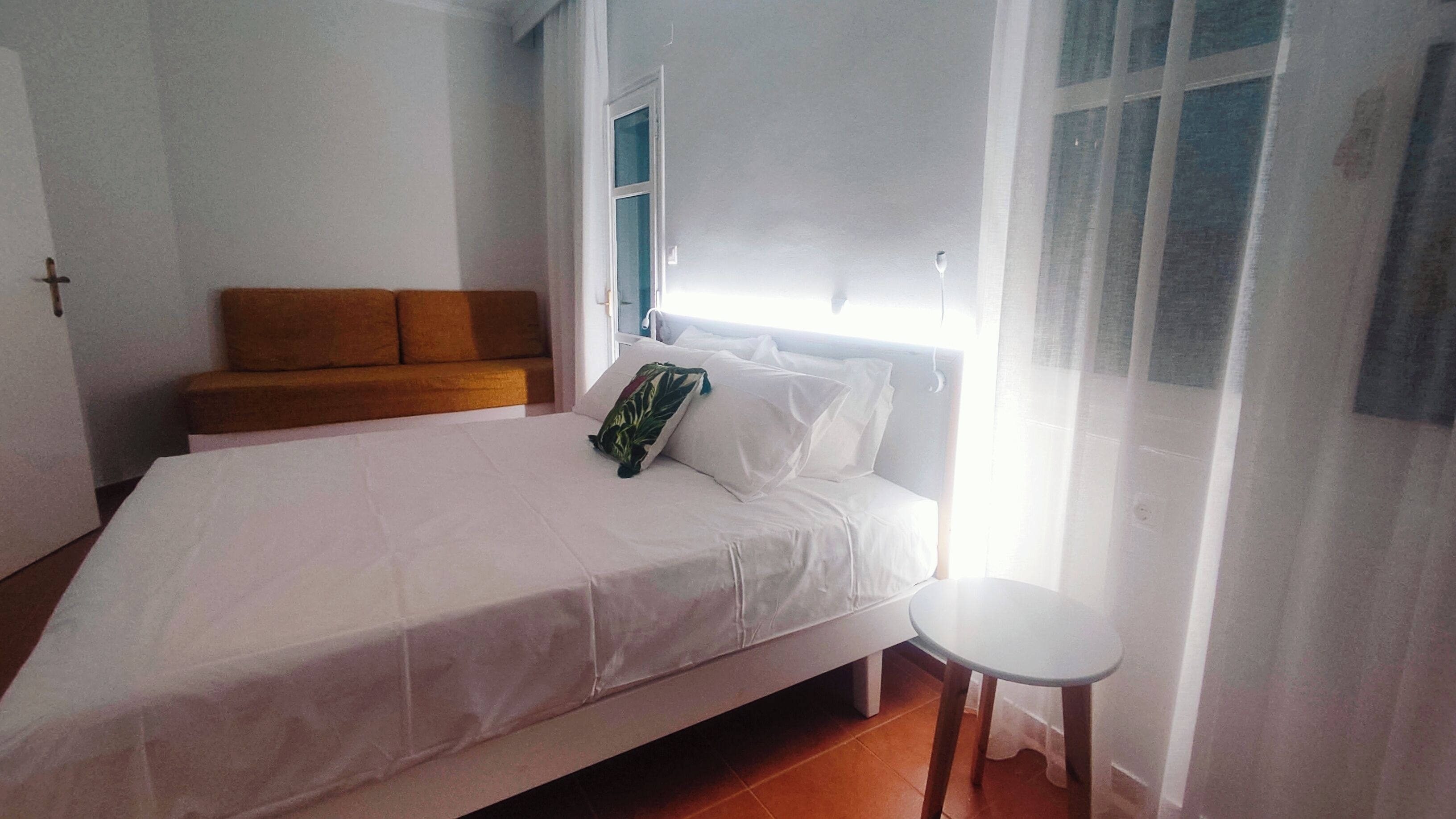 A bedroom with a bed, a small table, a sofa, and a window with sheer curtains.