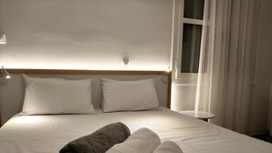 A neatly made bed with white and gray towels, a small bedside table, and a window with curtains. - Tarmaros - Families & Groups (Sissi)