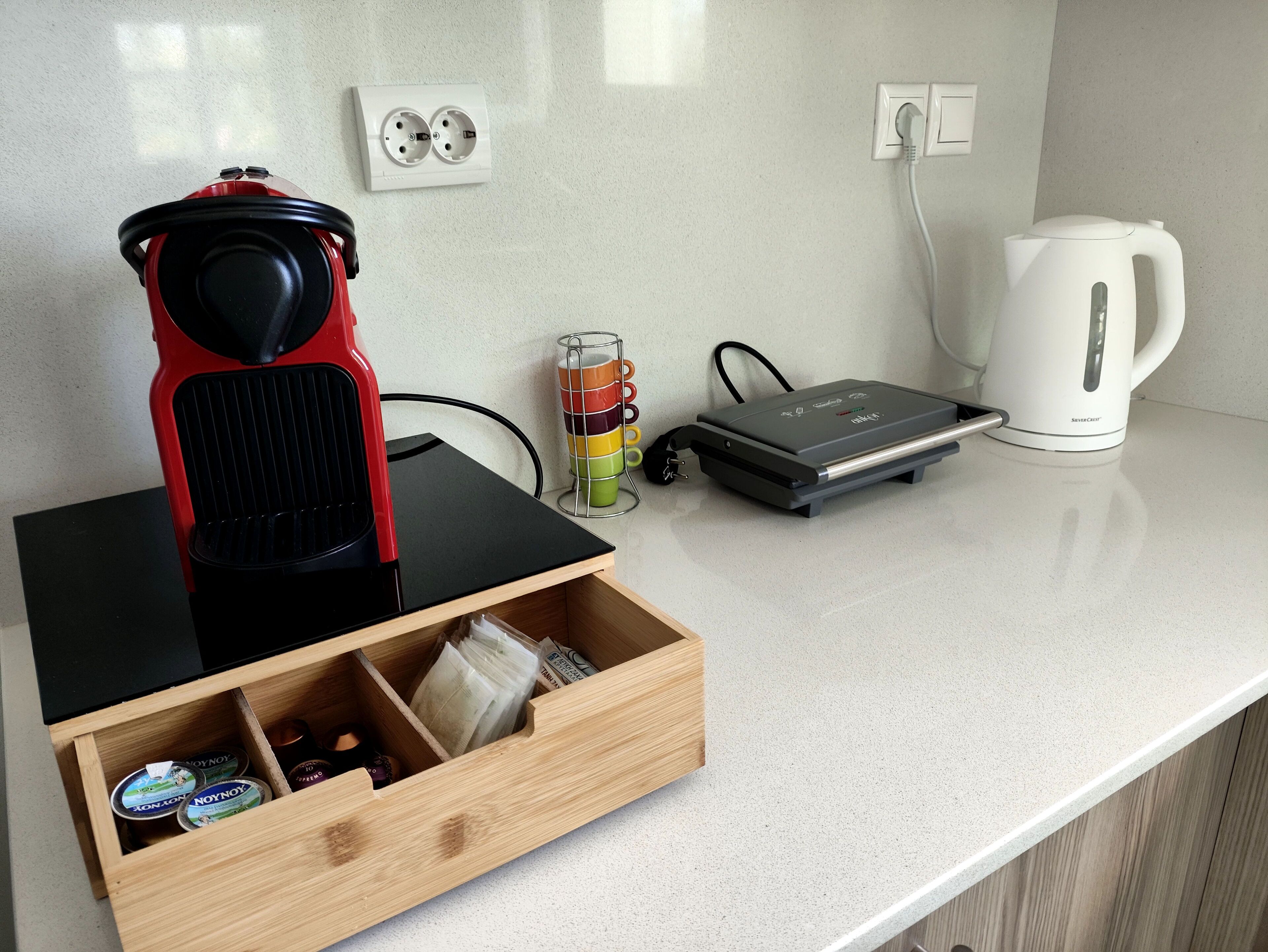 Un plan de travail de cuisine avec une machine à café rouge, une bouilloire blanche, un radiateur électrique portatif et un tiroir contenant des produits alimentaires.