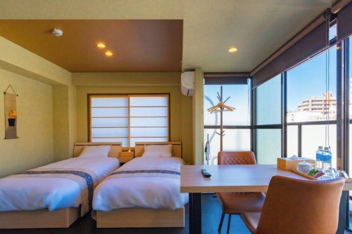 Quarto de hotel com duas camas, uma mesa, cadeiras, janela com vista para edifícios e um ventilador de teto.