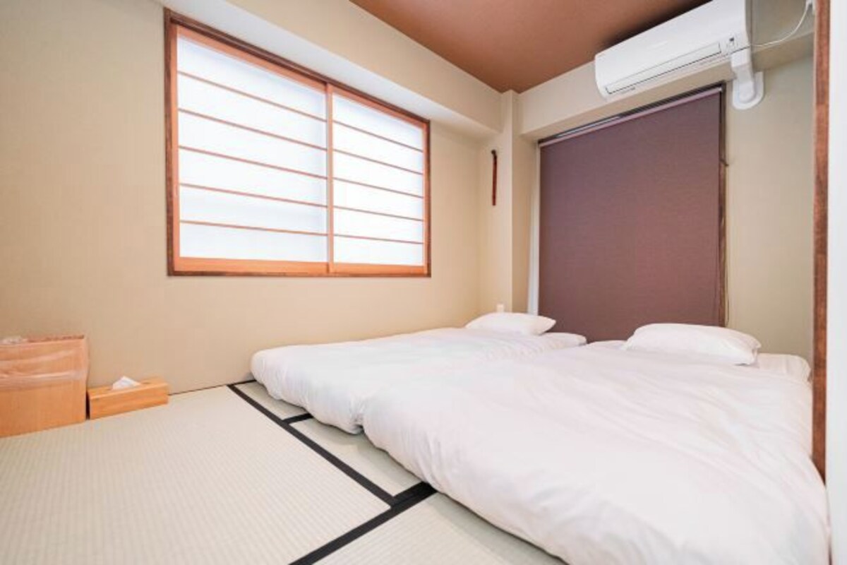 Une pièce avec un sol recouvert de tatamis, un lit avec une literie blanche, une porte coulissante et une fenêtre avec des stores en bois.