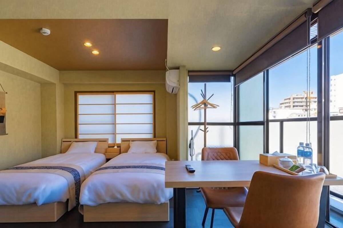 Quarto de hotel com duas camas, uma mesa com cadeiras, uma janela com vista e um ventilador de teto.