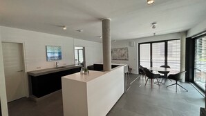 Een moderne keuken met een centraal kookeiland, een eethoek met een ronde tafel en stoelen, en grote ramen met zonwering.